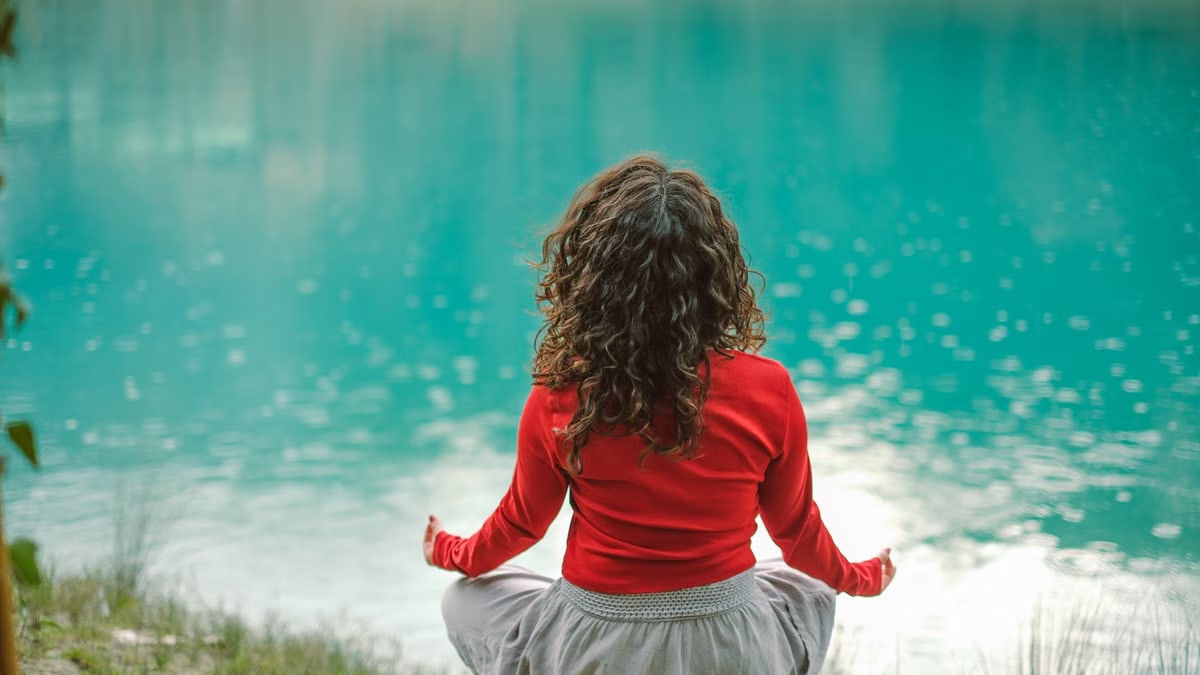 A woman in red meditates by a tranquil blue lake, reflecting calm and serenity.